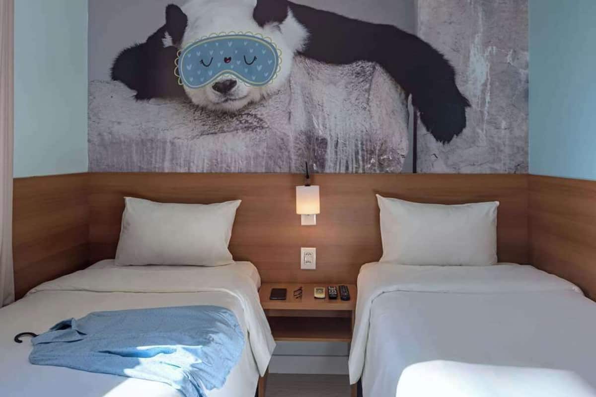 A hotel room with two beds, a wooden headboard, a nightstand with a lamp, and a wall mural of a panda.
