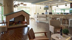 A modern hotel lobby with a wooden play area, comfortable seating, a bar, and a television.