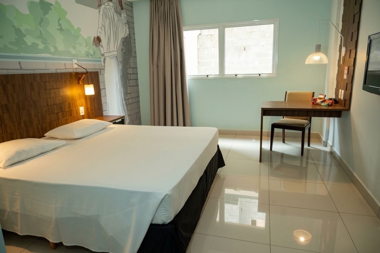 A hotel room with a bed, a desk, a chair, a window, and a wall-mounted lamp.