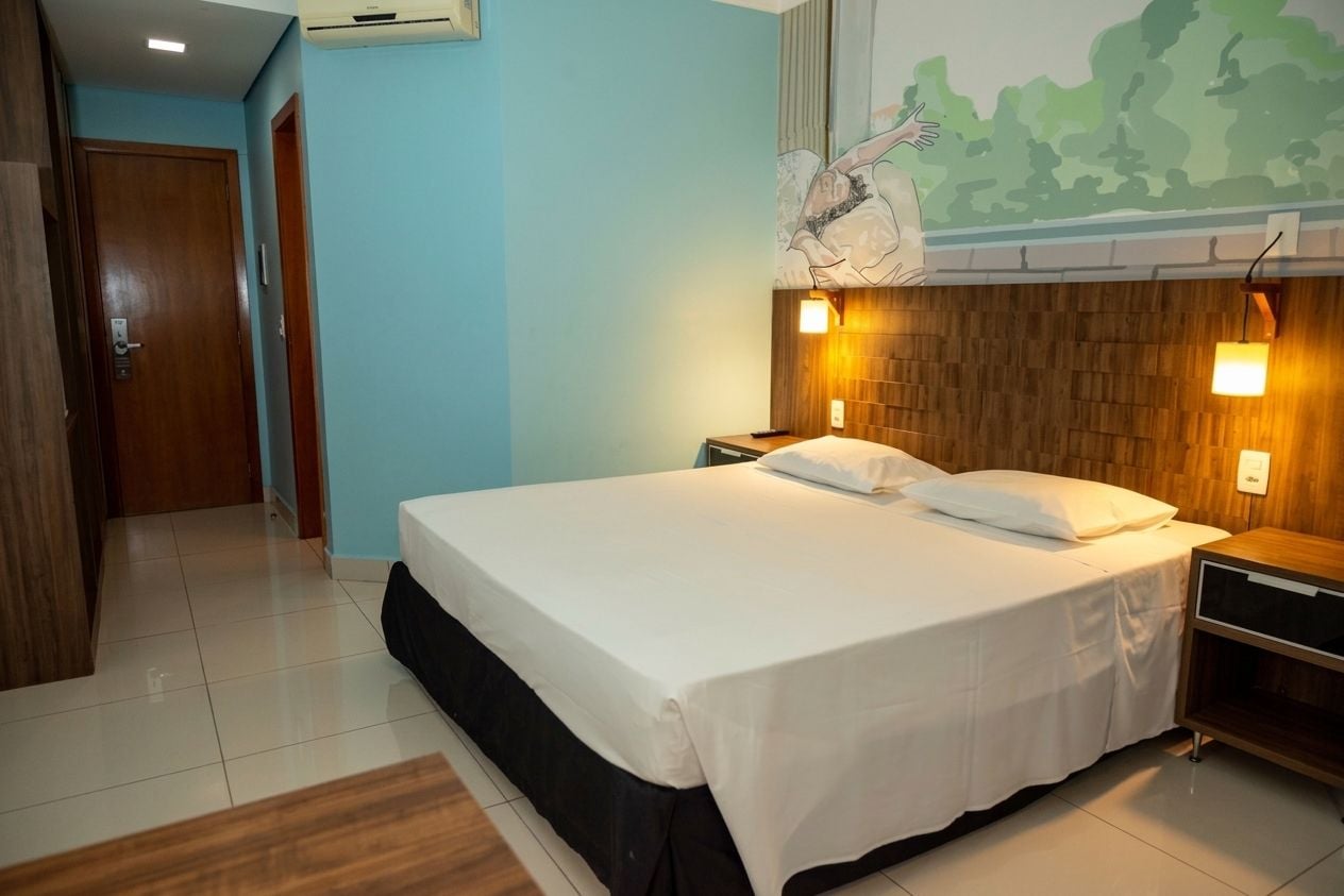 A hotel room with a bed, a nightstand, a wall-mounted air conditioner, and a door.
