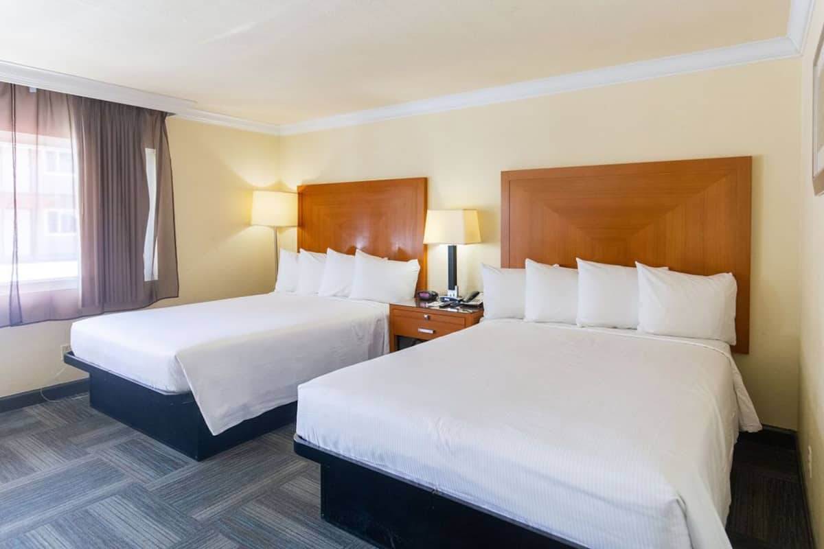 Habitación de hotel con dos camas, cada una con cabecero de madera, ropa de cama blanca y mesita de noche con lámpara.