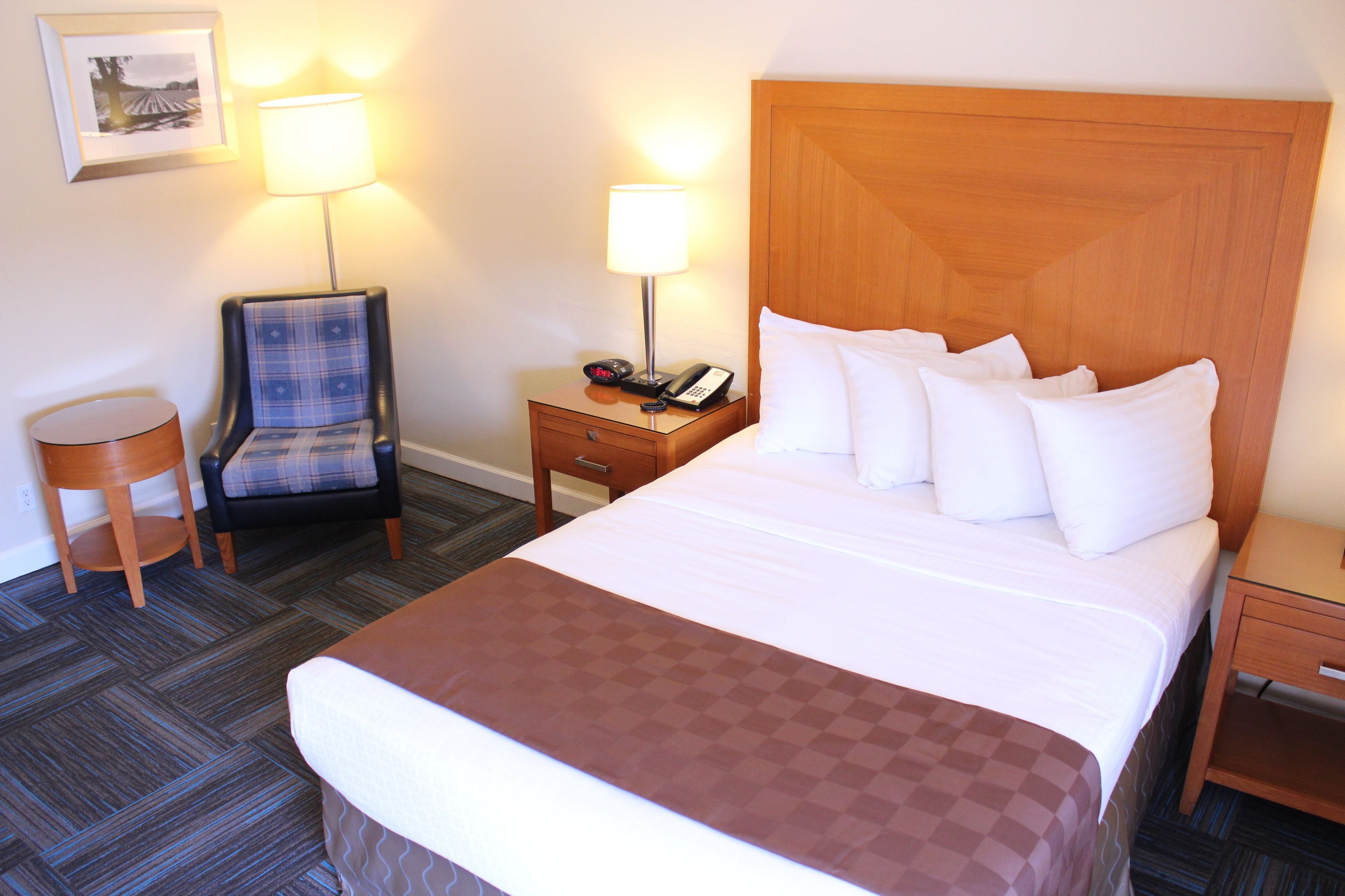 A hotel room with a large bed, a wooden headboard, a nightstand with a lamp, a chair, and a small table.