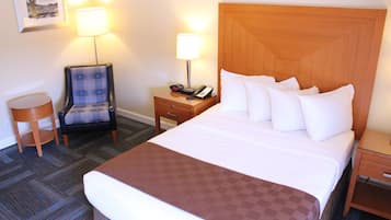 A hotel room with a large bed, a wooden headboard, a nightstand with a lamp, a chair, and a small table.