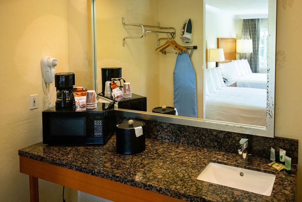 Baño de hotel con encimera de granito, lavabo, espejo, secador de pelo, cafetera y artículos de aseo.