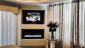 Una sala de estar con un televisor de pantalla plana montado en la pared, una chimenea debajo y una mesa de vidrio con un jarrón de flores.