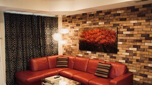 Una sala de estar con un sofá modular, una mesa de centro de vidrio, una cortina estampada y una pared de ladrillo con un cuadro.