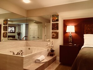 A modern bathroom with a large bathtub, a mirror, a vanity with a lamp, and a bed.