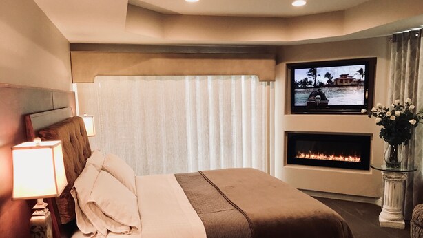 Habitación de hotel con cama, televisor montado en la pared, chimenea y mesita de noche con lámpara.