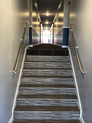 A modern hotel hallway with a patterned carpet, metallic handrails, and a view of the exit at the end. - Relax Inn Motel (Los Angeles)