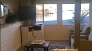 A living room with a leather sofa, a coffee table, and a TV on the wall. There are windows with a view of the outside.