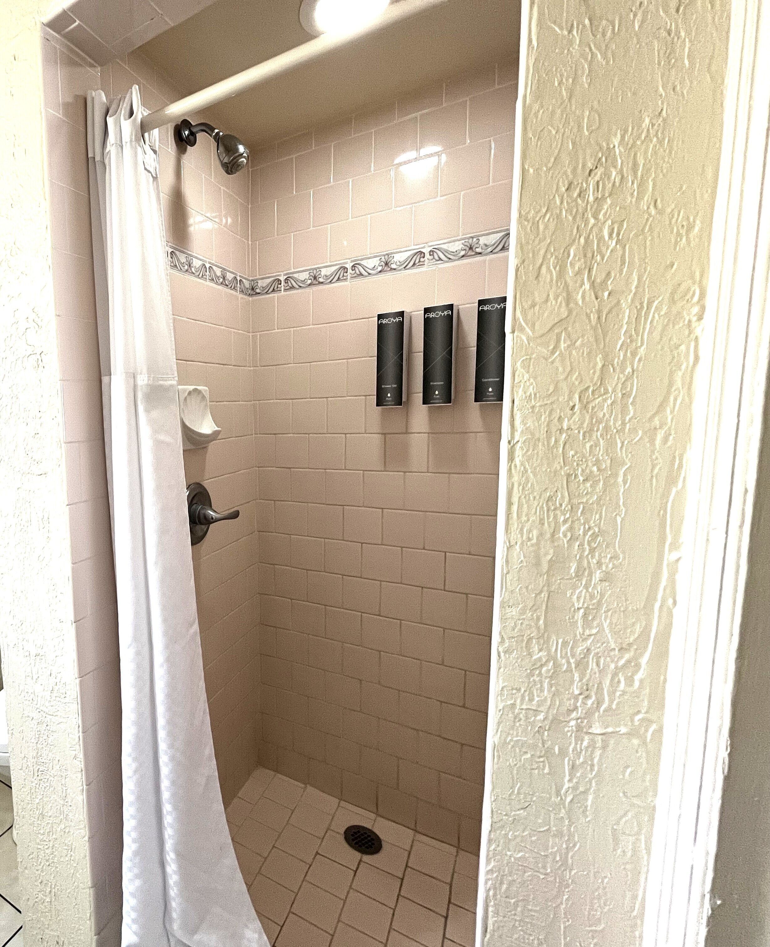Ducha con azulejos color beige, una cortina blanca y tres dispensadores negros en la pared.