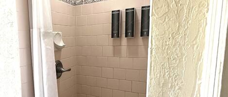 Ducha con azulejos color beige, una cortina blanca y tres dispensadores negros en la pared.
