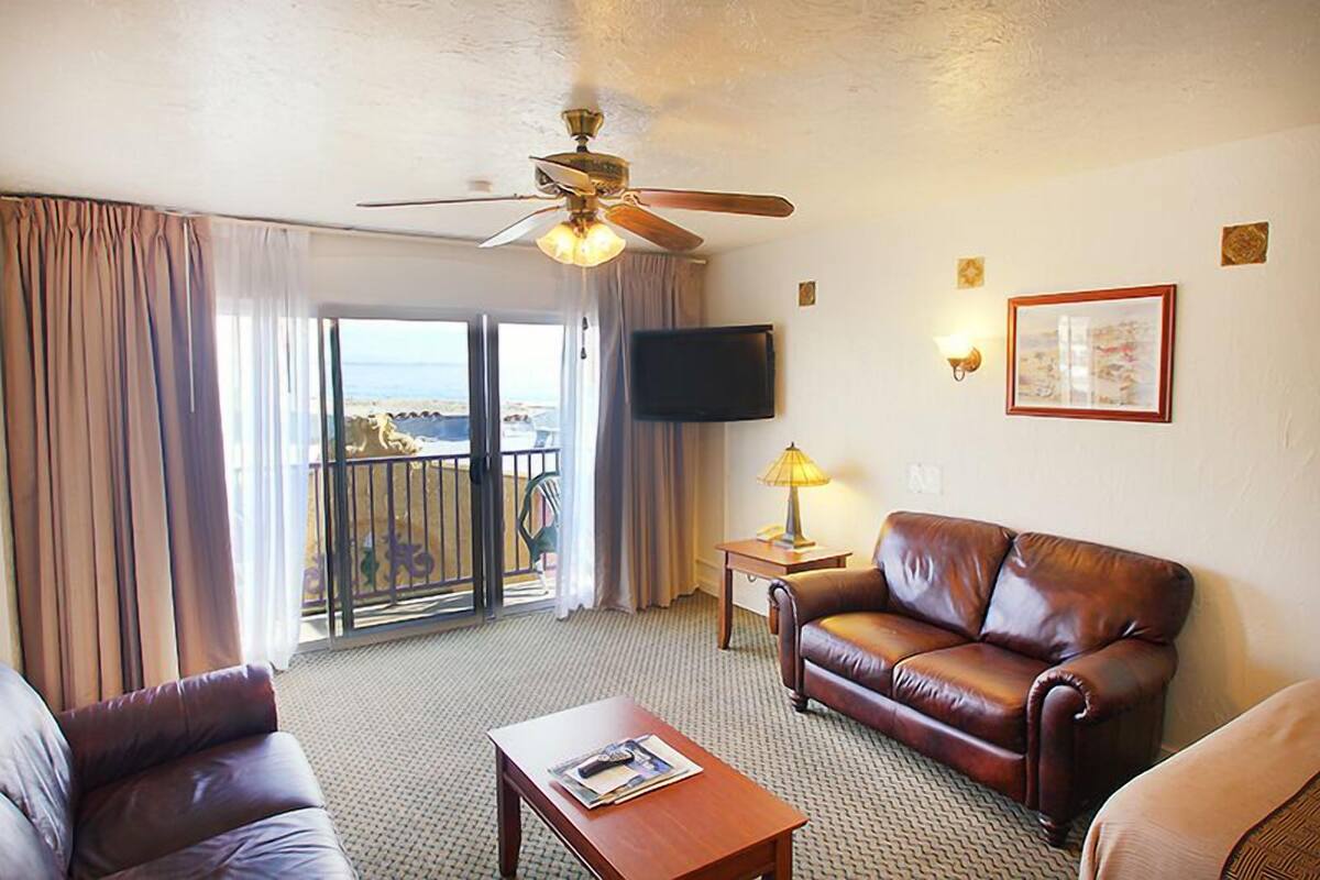 A hotel room with a leather sofa, a coffee table, a ceiling fan, a TV, a balcony with a view, and a framed picture on the wall.