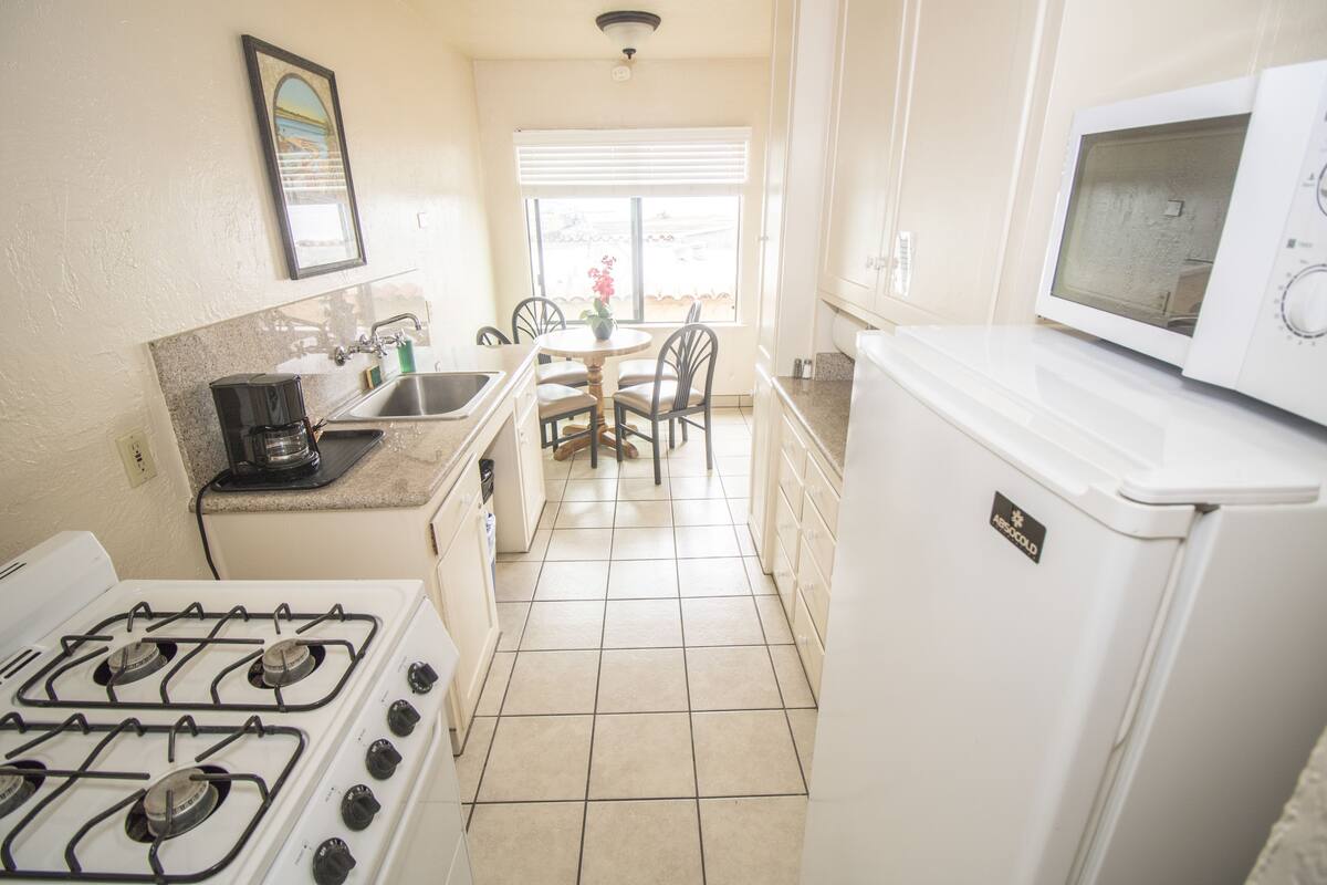 A small kitchen with a gas stove, sink, and a microwave. There is a window with blinds, a chair, and a table.