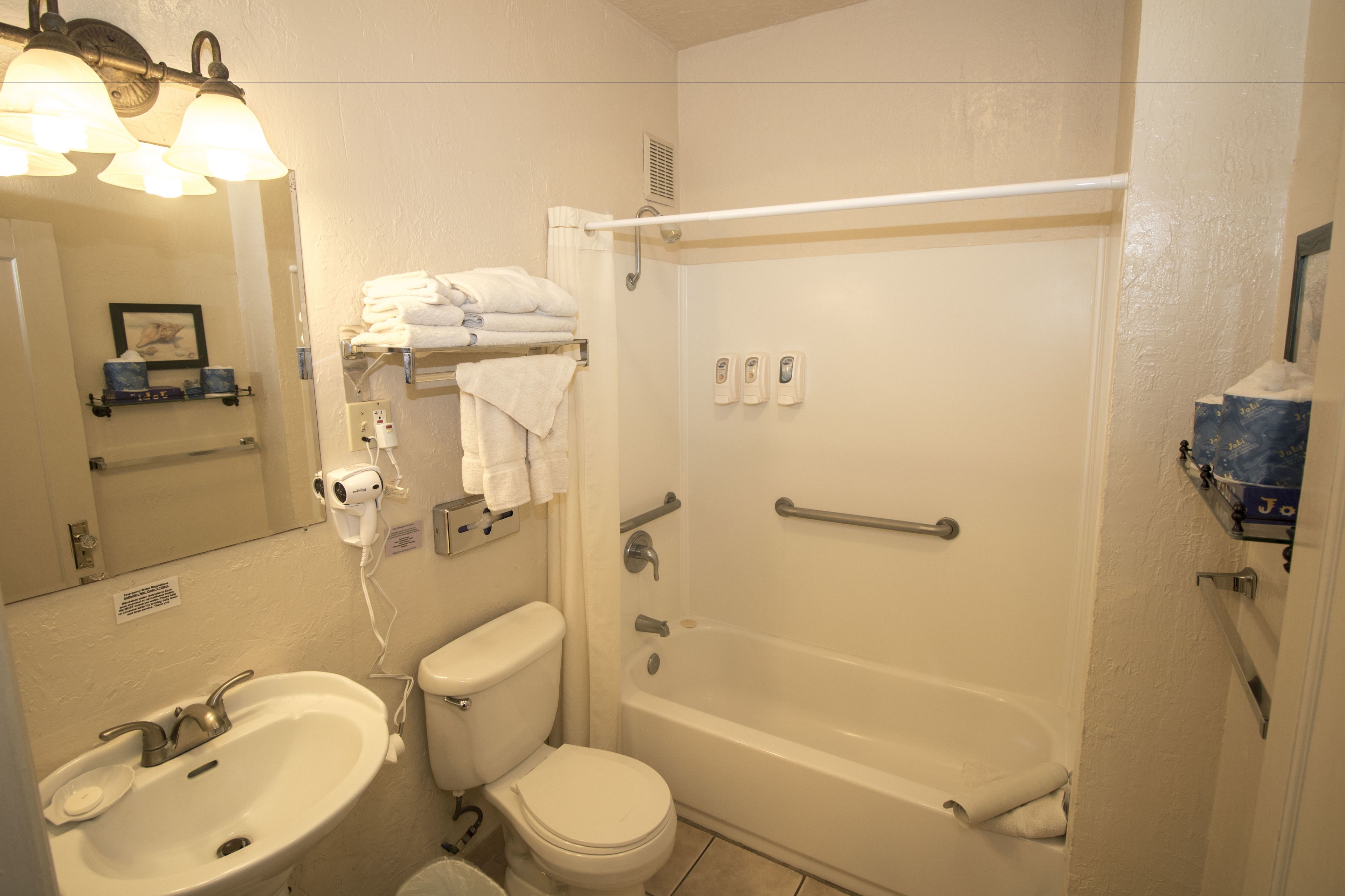 A bathroom with a sink, toilet, and bathtub. There are towels stacked on a shelf, a hand dryer, and a mirror.