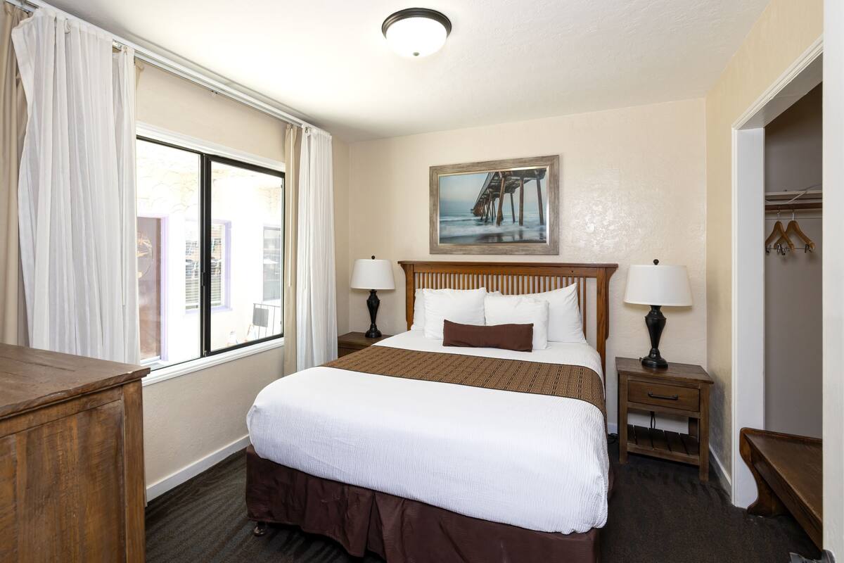 A hotel room with a large bed, two bedside tables with lamps, a painting on the wall, and a window with curtains.
