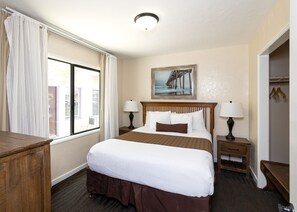 A hotel room with a large bed, two bedside tables with lamps, a painting on the wall, and a window with curtains. - Capitola Venetian Vacation Rentals (Capitola)