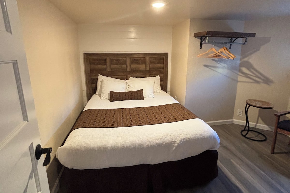 A small, narrow hotel room with a single bed, a wooden headboard, a small table with a chair, and a coat rack with hangers.
