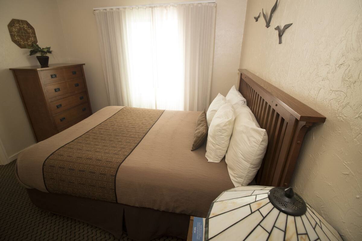 A neatly made bed with a patterned bedspread and pillows, a wooden nightstand with a lamp, a dresser with drawers, and a window with sheer curtains.