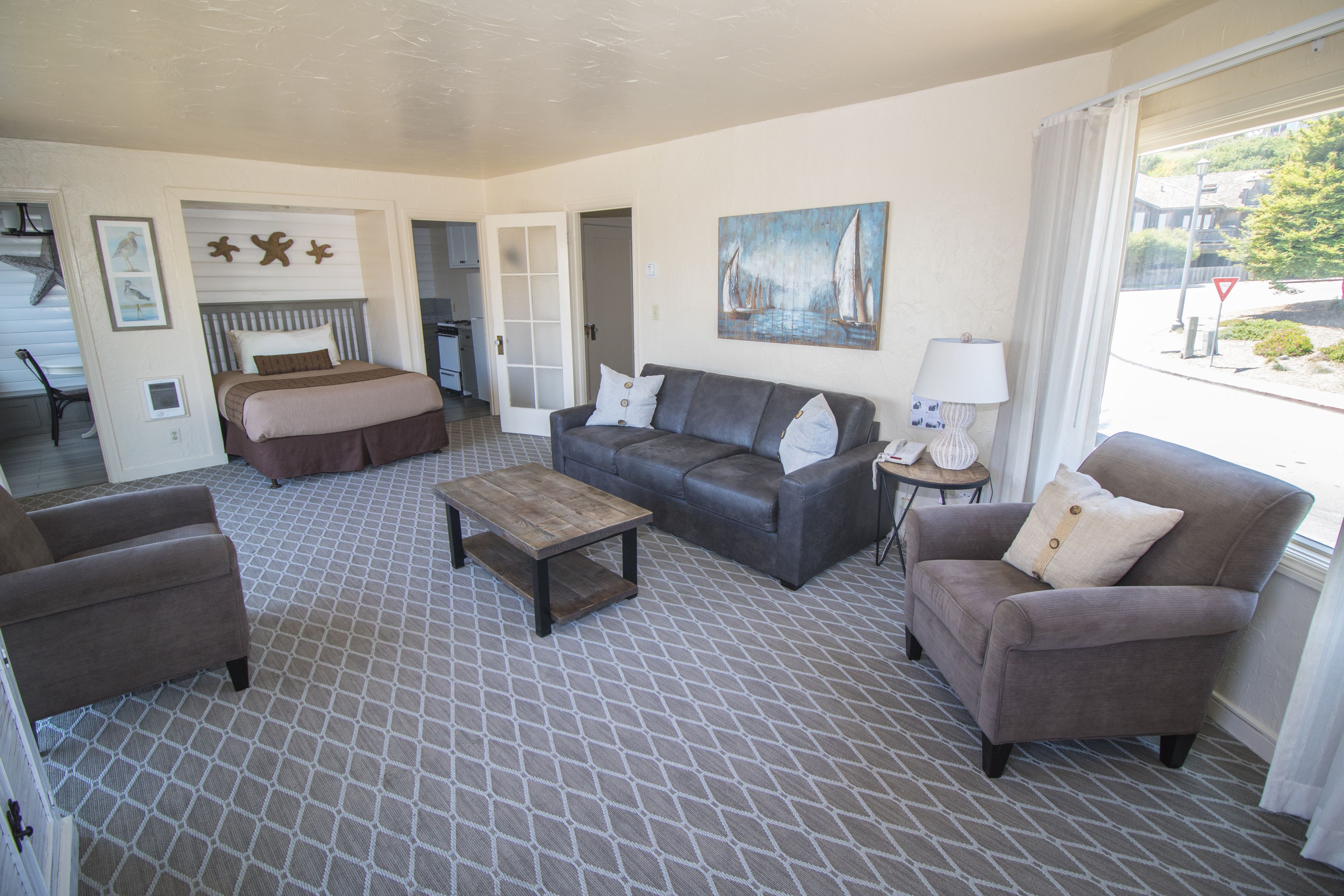 A hotel room with a bed, a sofa, a coffee table, and a chair. There is a painting on the wall and a window with curtains.