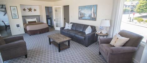 A hotel room with a bed, a sofa, a coffee table, and a chair. There is a painting on the wall and a window with curtains.