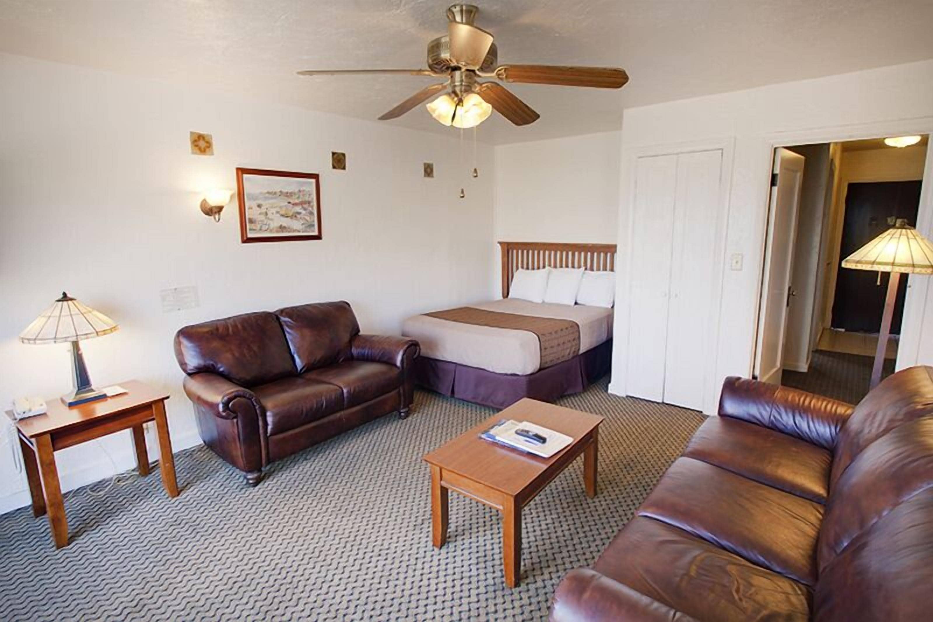 Une chambre d’hôtel avec un canapé en cuir marron, une table basse en bois, un lit avec une tête de lit en bois, un ventilateur de plafond et une lampe sur une table d’appoint.