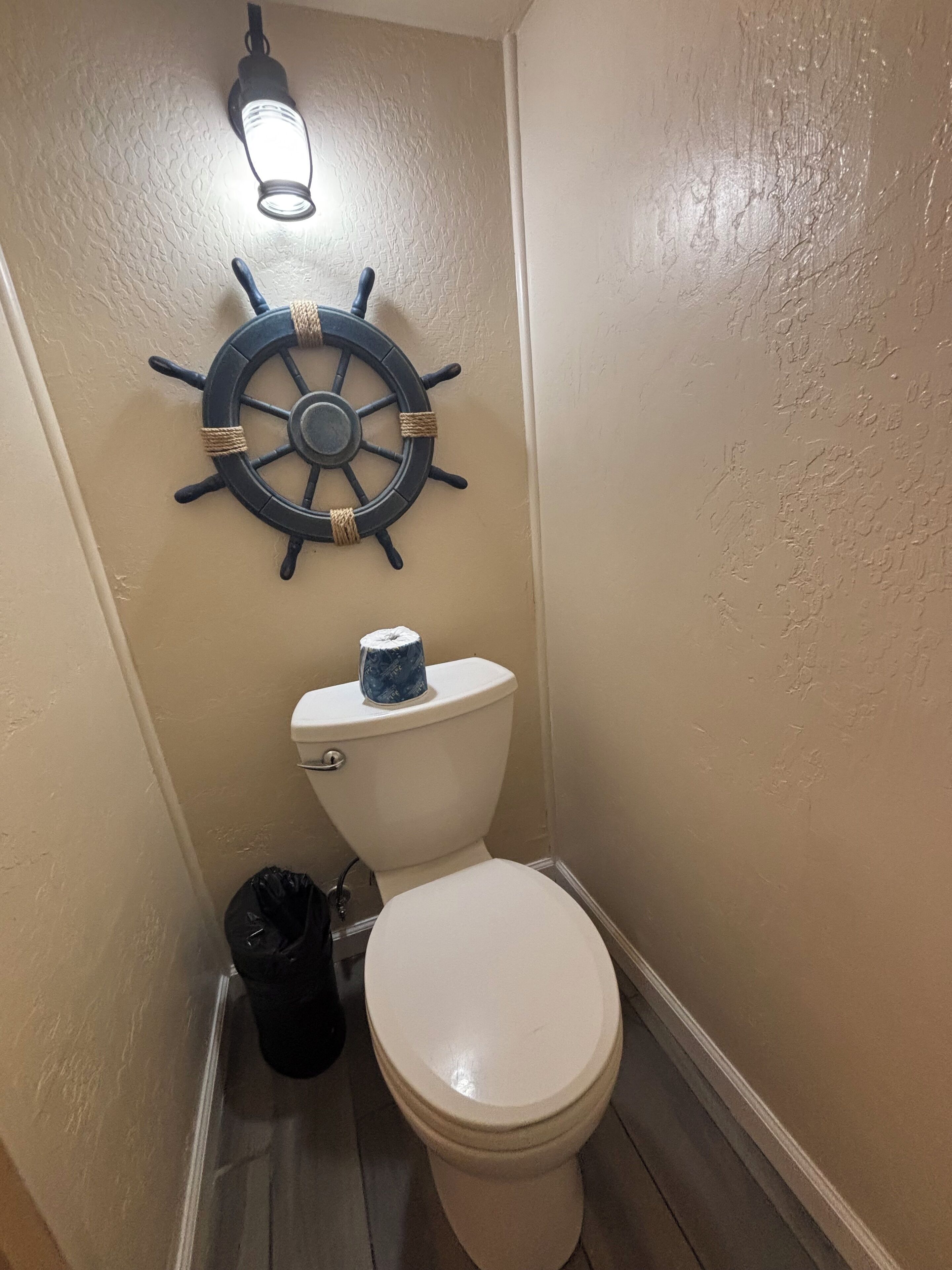 A bathroom with a toilet, a ship's wheel decoration, and a trash bin.