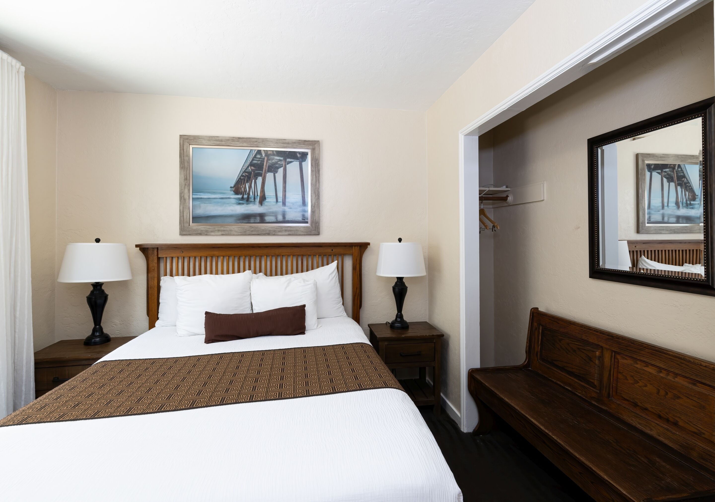 A hotel room with a large bed, two bedside tables with lamps, a wooden bench, a framed picture on the wall, and a mirror reflecting the bed.