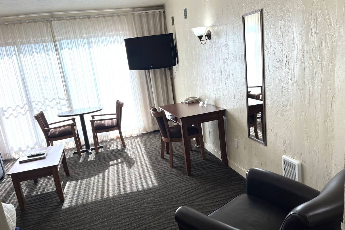 A hotel room with a flat-screen TV mounted on the wall, a desk with a telephone, two chairs, a small table, and a large mirror.