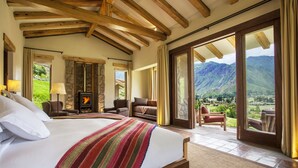 A spacious bedroom with a large bed, a stone fireplace, and a view of a mountainous landscape through a sliding glass door. - Inkaterra Hacienda Urubamba (Huayllabamba)