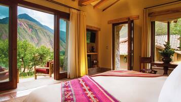 A bedroom with a large bed, wooden beams, a view of mountains, and a balcony with a chair.