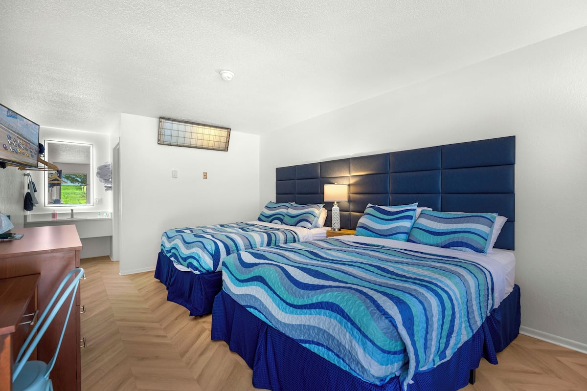 A bedroom with two beds, a wooden dresser, a window with a view, and a wall-mounted lamp.