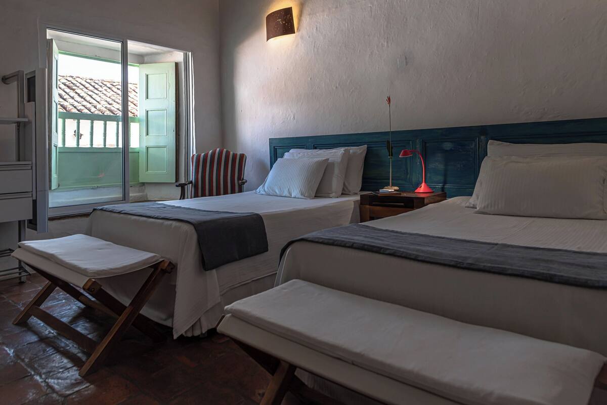 A room with two beds, a bench, a nightstand, a lamp, and a window with a view of a building.