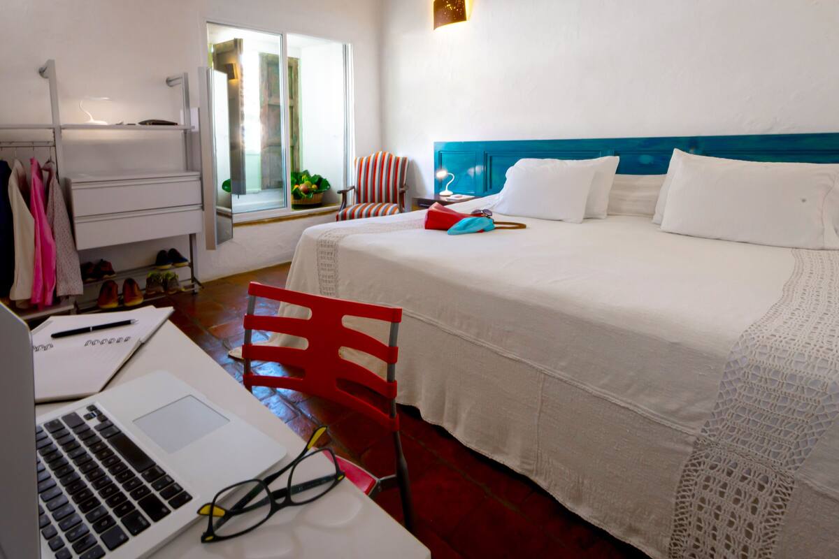 A hotel room with a bed, a desk with a laptop, a chair, a window with a view, and a door.