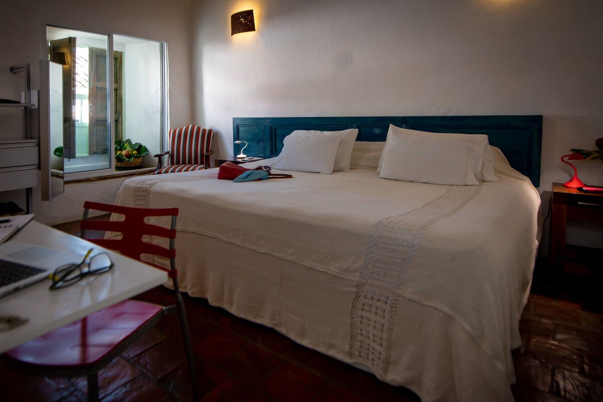 A hotel room with a bed, a desk with a laptop, a chair, and a window with a view of the outside.