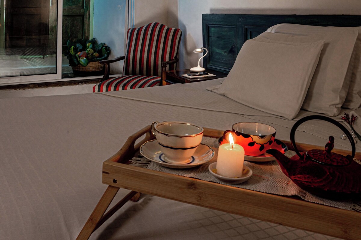 A bed with a bedside table, a chair, a tray with a candle, teapot, and cups.