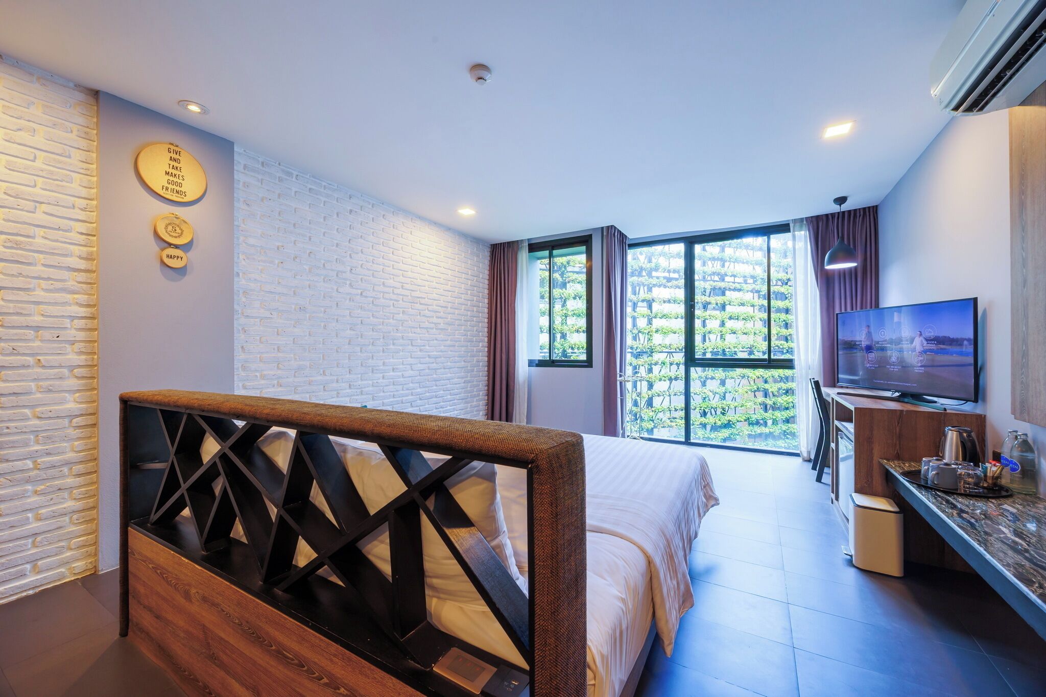 A modern hotel room with a bed, a TV, and a view of greenery outside.
