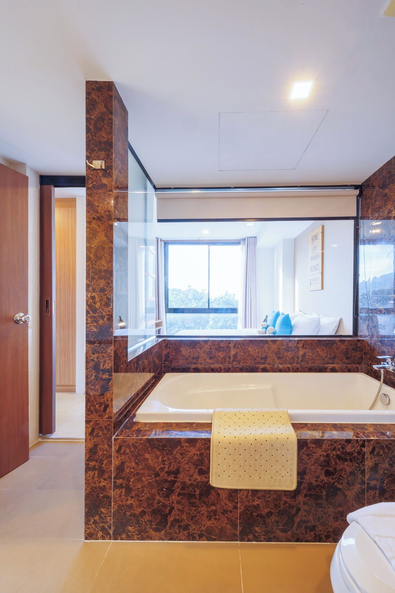 A modern bathroom with a large bathtub, a mirror, and a view of a room with a bed and window.