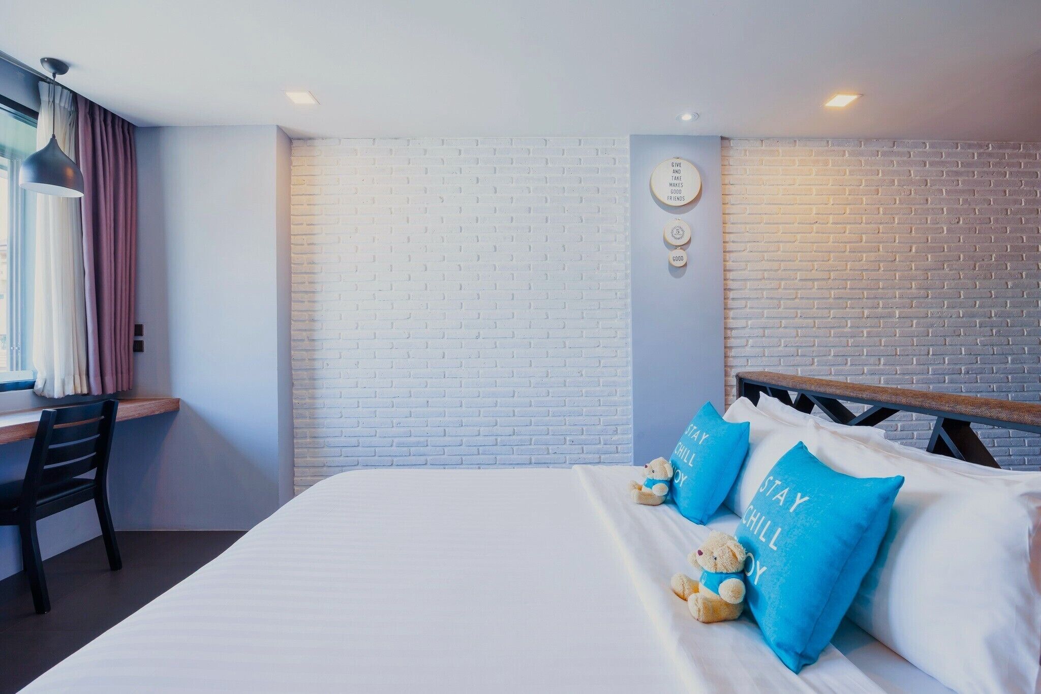 A hotel room with a white brick wall, a desk with a chair, a bed with blue pillows, and a teddy bear on the bed.