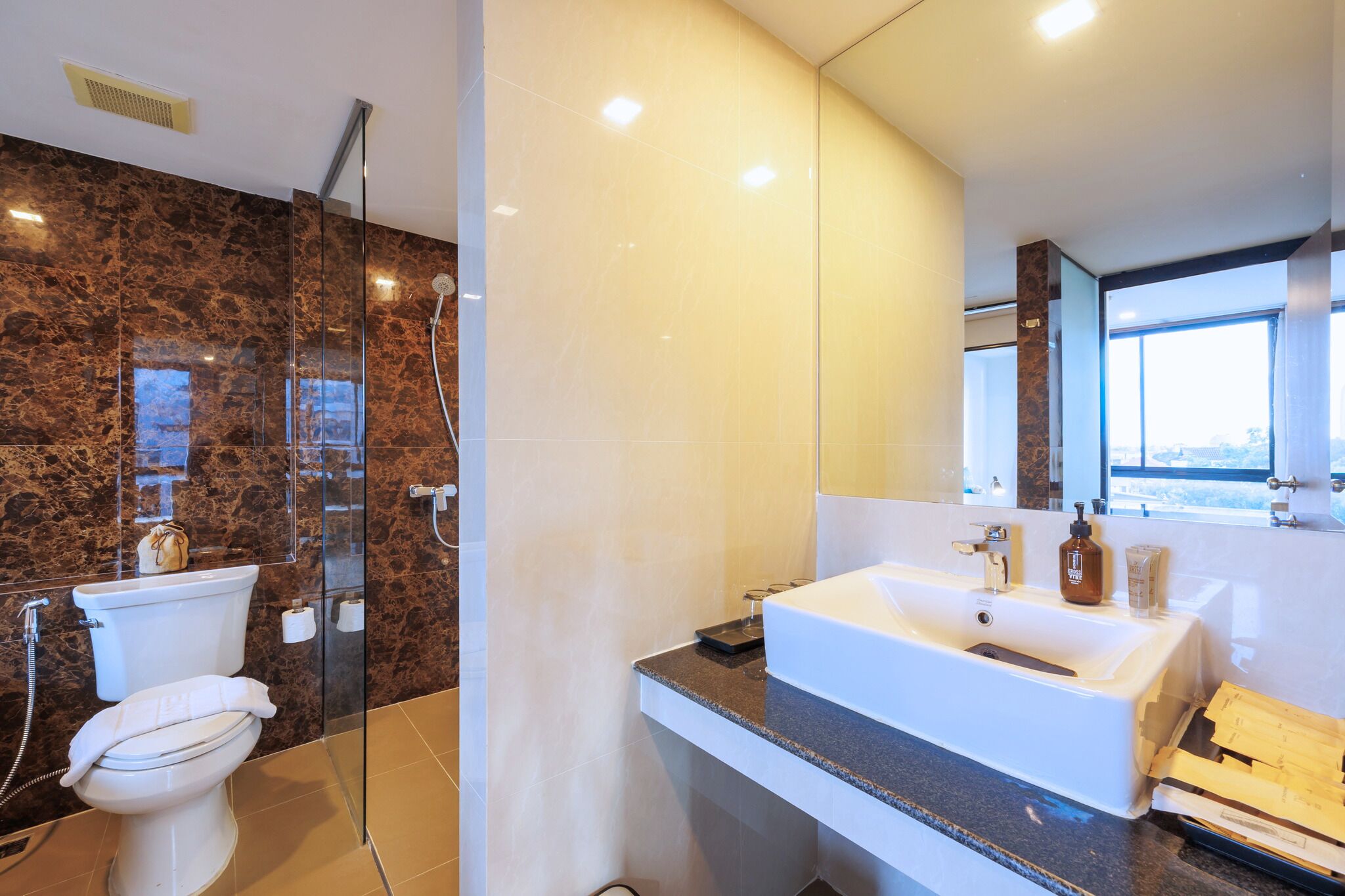 A modern bathroom with a glass-enclosed shower, a white toilet, a large sink with a mirror, and a window providing natural light.