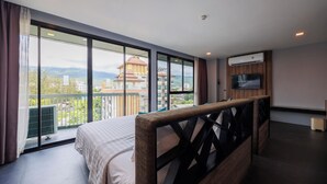 A hotel room with a large bed, a TV mounted on the wall, and a balcony with a view of buildings and mountains. - Cross Vibe Chiang Mai Decem (Chiang Mai)