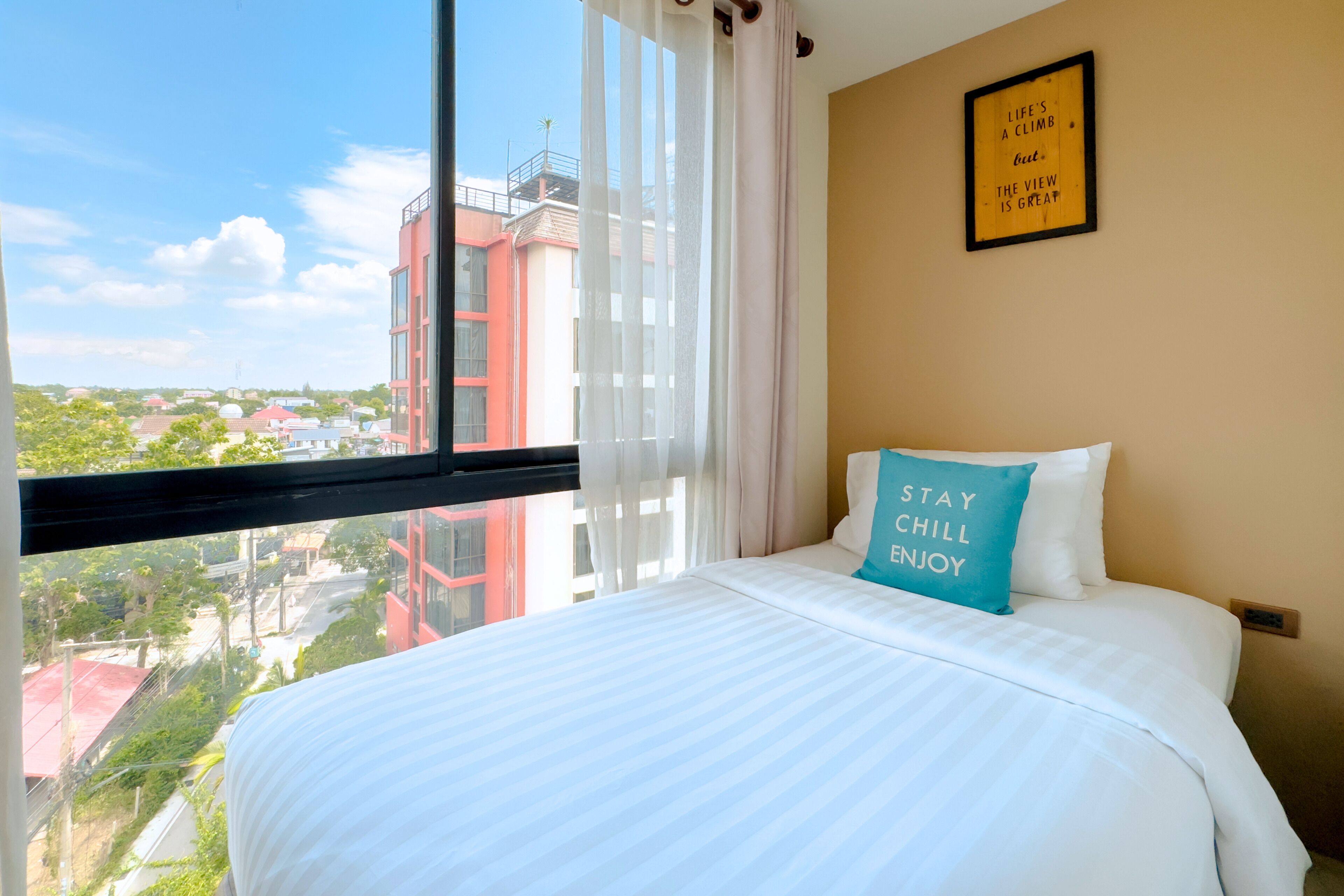A hotel room with a large window, a bed with white bedding, a teal pillow with text, and a framed quote on the wall.