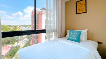 A hotel room with a large window, a bed with white bedding, a teal pillow with text, and a framed quote on the wall.