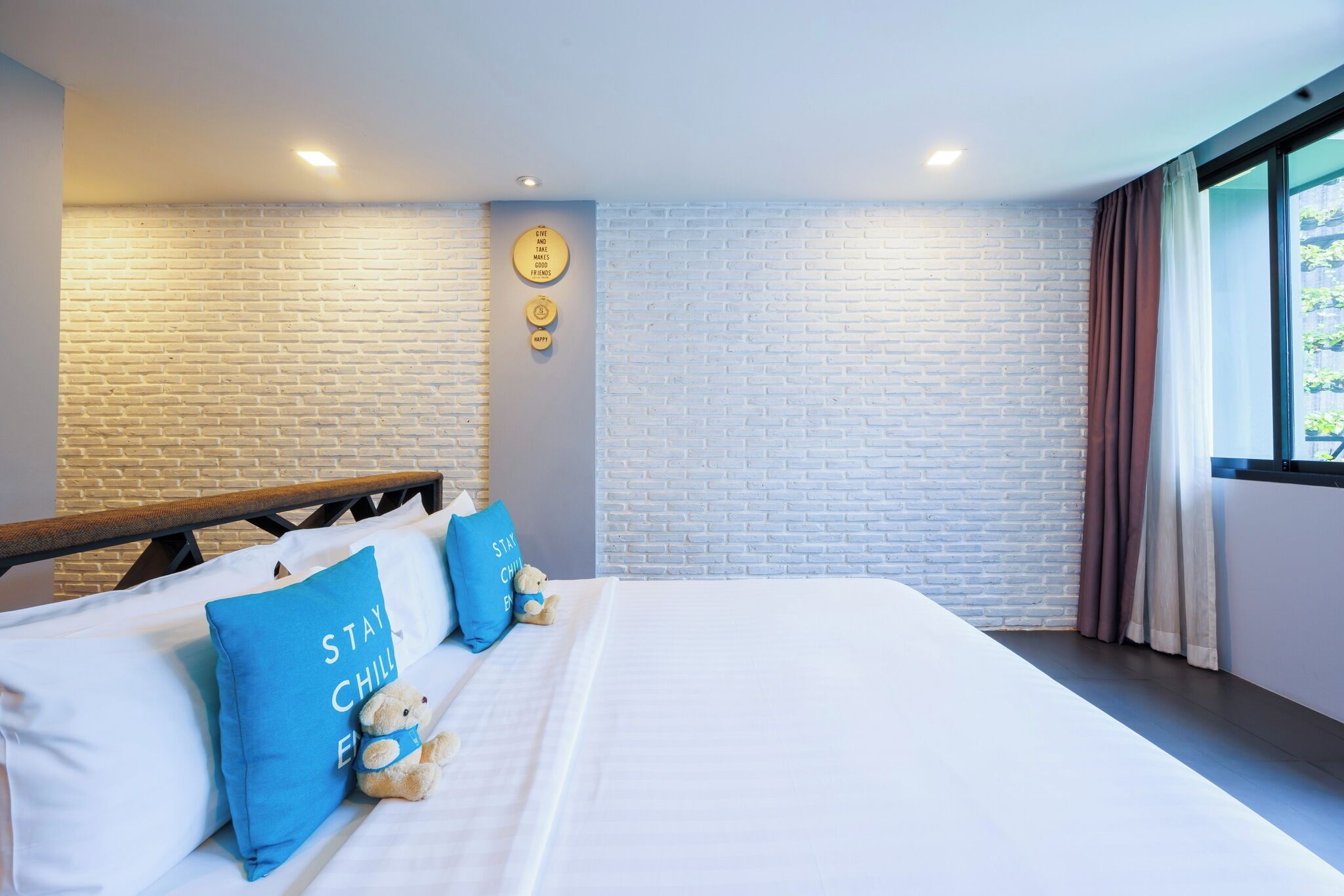 Una habitación de hotel moderna con pared de ladrillo, cama con sábanas blancas, dos almohadas azules, un oso de peluche y una ventana con cortinas.