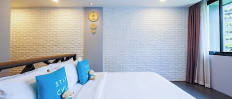A modern hotel room with a brick-patterned wall, a bed with white linens, two blue pillows, a teddy bear, and a window with curtains.