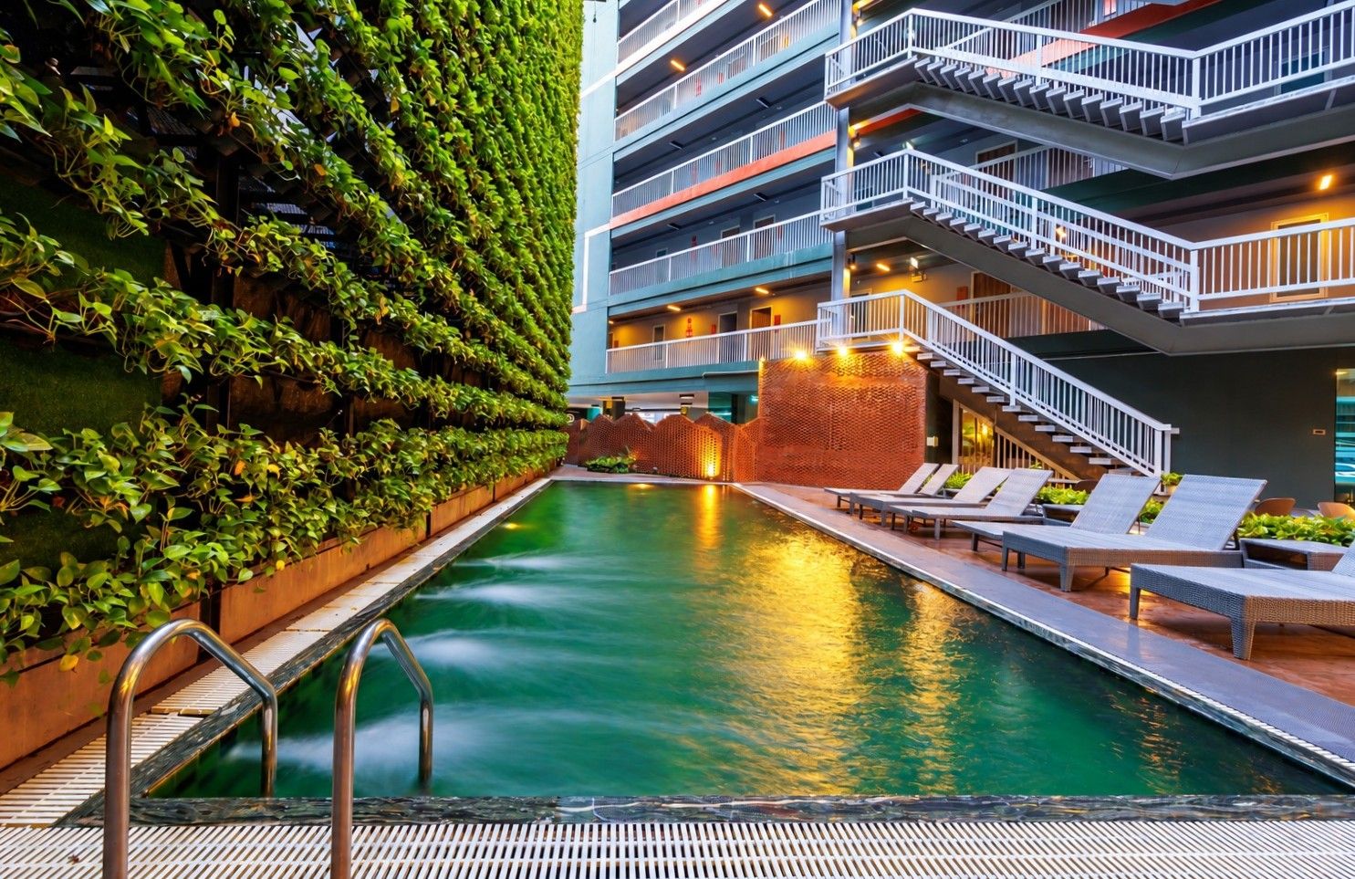 A swimming pool with a metal ladder, surrounded by lounge chairs and a building with a green wall and multiple balconies.