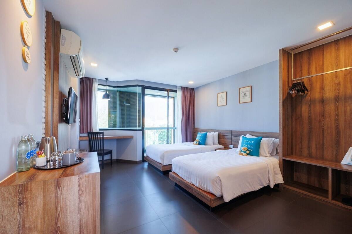A hotel room with two beds, a desk with a chair, a large window, and a wooden wardrobe.