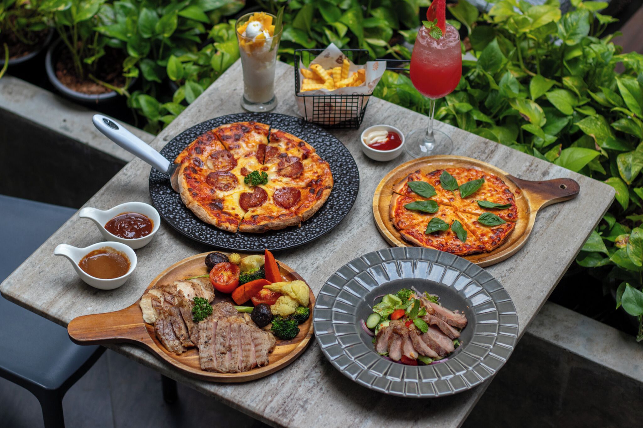 Une table dressée avec deux pizzas, un plat de viande, une salade et des condiments.