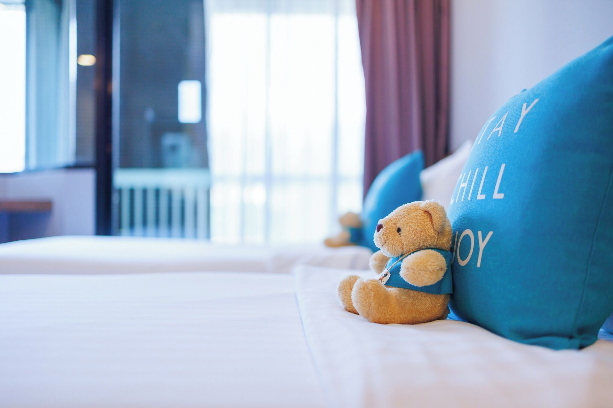 A bed with a teddy bear and pillows with text, in a room with a window and curtains.