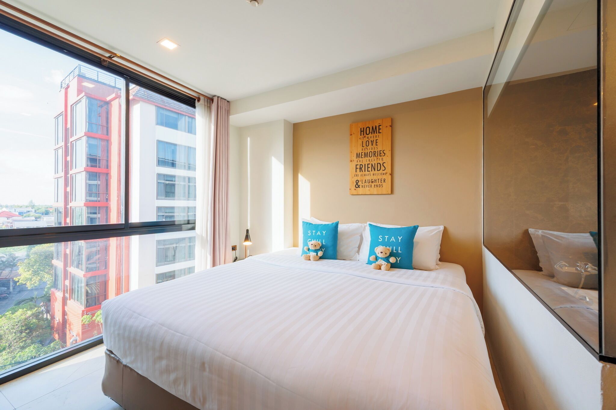 A hotel room with a large bed, a view of buildings through a window, a wall with a motivational quote, and a bedside table with a lamp.